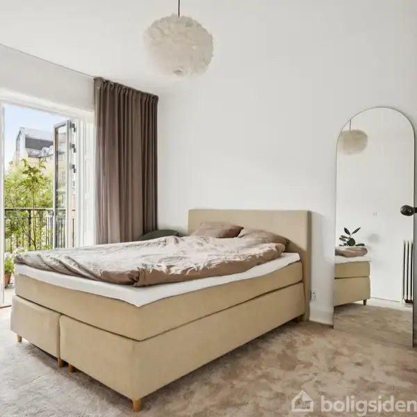 Soveværelse med dobbeltseng står foran en åben altandør. Langt spejl og potteplante ses ved siden af sengen. Beige tæppe og hvid loftslampe dominerer rummet, der har neutrale farver.
