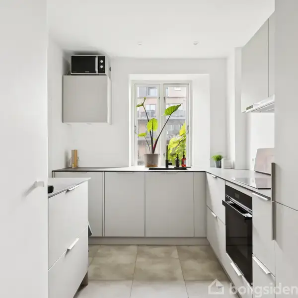 Et lyst, moderne køkken med hvide skabe og integrerede apparater. En vindueskarm viser planter, der giver farve. Flisegulv skaber et rent og minimalistisk udseende.