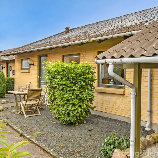Et gult murstenshus med en tagrende, omgivet af en gårdhave med hæk og fliser. Et lille bord med to stole står ved siden af en grøn busk i haven.