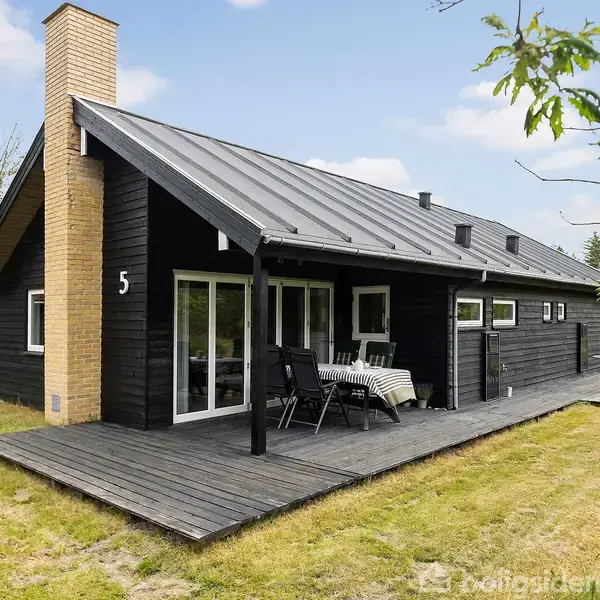 Sort træhus med skråt tag, terrasse og skorsten. På terrassen står et bord med stole. Græsplæne omgiver huset, og et skovområde kan ses i baggrunden. Tallet "5" er på huset.