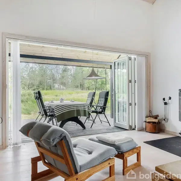 En moderne stue med en pejs til højre, åbne foldedøre til en terrasse med bord og stole, omgivet af en grøn have.