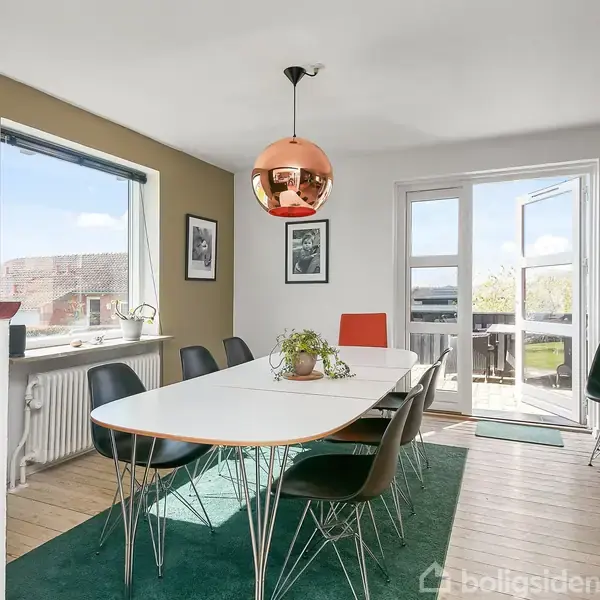 Spisebord med sorte stole står på en grøn måtte. En kobberlampe hænger over bordet. Rummet har lyse trægulve, et vindue og en åben dør til en altan. Planter og billeder dekorerer rummet.