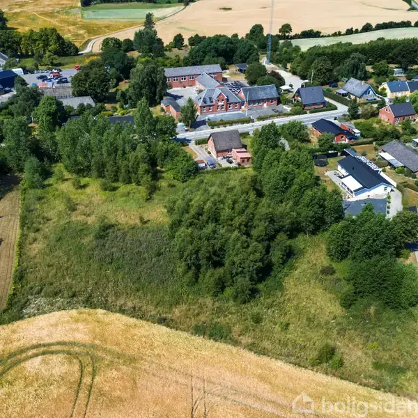 Luftfoto af landsbyområde, der viser en klynge af træer omkring åbne marker med spredte huse og en hovedvej. Baggrunden består af store marker og skovområder.