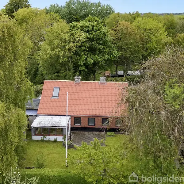 Hus med rødt tag står i en frodig have omgivet af tæt vegetation og store træer, i et landligt område uden andre bygninger tæt på.