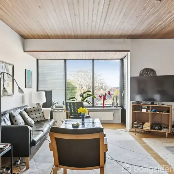 Stue med sofa, lænestol og lamper, hvor vinduet bagved viser udsigt. Et TV står på reol til højre med en rød stol ved siden af. Værelset har trægulv og -loft.