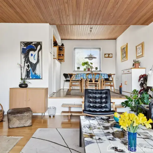 Stue med moderne møblering; en sort lænestol og sofa foran et sofabord med gule blomster; spisestue i baggrunden; kunst på væggene og træloft foroven.