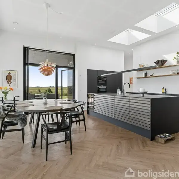 Et moderne køkken-alrum med et rundt spisebord og sorte stole. Køkkenøen er mørk, omgivet af en lys, rummelig indretning med store vinduer og udsigt til det grønne landskab.
