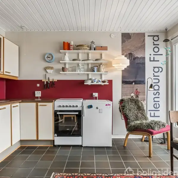 Køkken med hvide skabe og en rød bagvæg. Komfur og opvaskemaskine til venstre; hylder med service ovenover. En stol med pelsdækken står ved bordet. Tekst: "Flensburg Fjord".