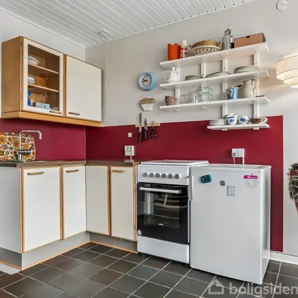 Køkkenet har et komfur ved siden af et lille køleskab. Åbne hylder og skabe over disken opbevarer tallerkener og glas. Væggen er rød, og der er en lampe.