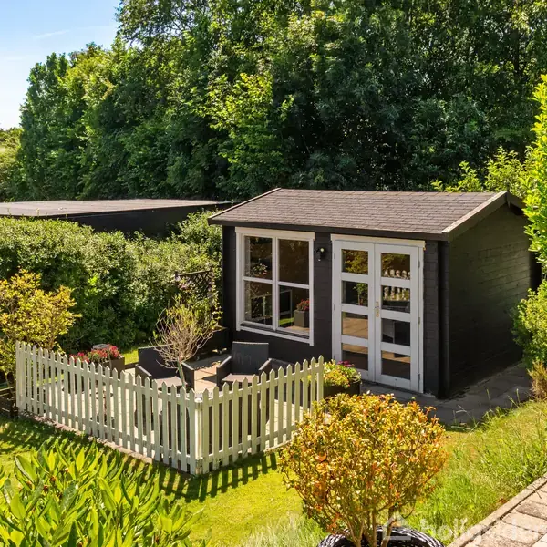 Et lille, sort havehus står omgivet af en hæk og grønne træer i en have. Foran huset er et hvidt stakit, og indeni er der havemøbler. Solen skinner.
