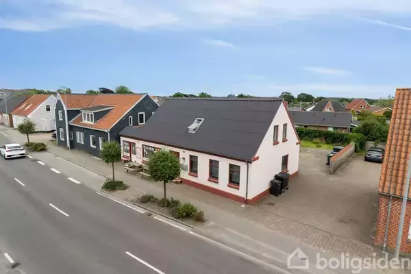 Et hvidt hus med rødt tag står langs en vej med fortov. Flere små træer og buske pryder foran huset. Andre huse kan ses i baggrunden under en blå himmel.