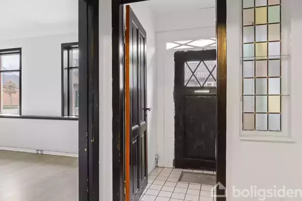 En åben sort hoveddør står på en hvid flisegulv, omgivet af hvide vægge med farvet glas og et tilstødende rum med store vinduer uden møbler.