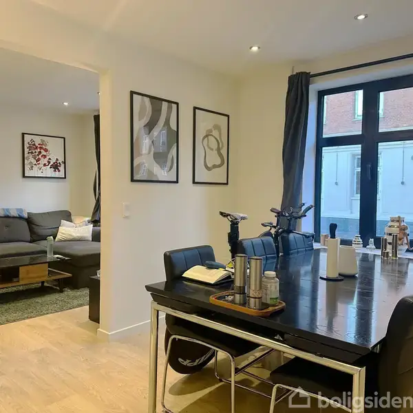 En moderne stue har en grå sofa mod en lys væg med to indrammede billeder. Foran vinduet er et sofabord, og til højre står et fladskærms-tv på en stander.