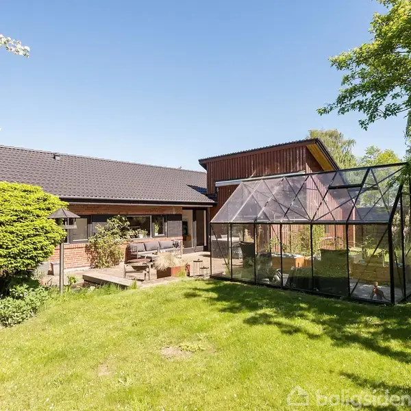 Glashus står stille i en frodig have ved siden af et moderne, murstenshus. Haven er grøn med buske og træer omkring, skaber en fredelig og naturlig atmosfære.