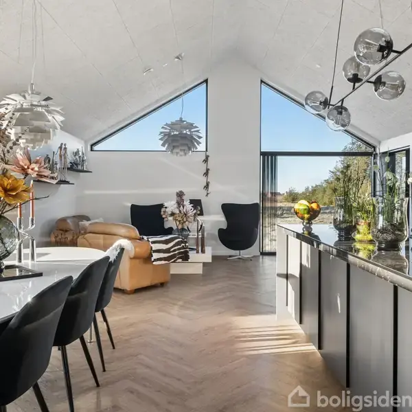 Spisebord med sorte stole, blomsterdekorationer, i en moderne stue med skrå lofter. Store vinduer lader lys ind, afslørende en lænestol og moderne lamper. Hus omkranset af natur.
