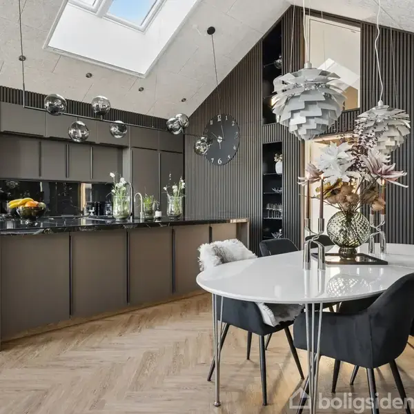 Køkken med grå skabe og en sort ovn i midten, omgivet af dekorative lamper. Bord dækket med blomsterarrangement og sorte stole under lysekrone. Store loftvinduer giver naturligt lys.