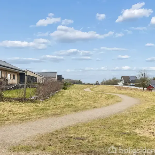 Hus står ved en grusvej, omgivet af græs og spredte buske, med flere huse i baggrunden under en blå himmel med spredte skyer.