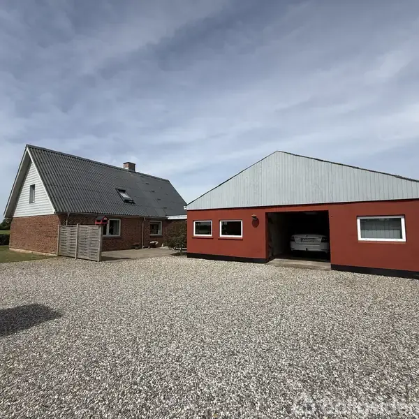 Et rødt hus med sort tag står ved siden af en separat garagebygning. En bil er parkeret i garagen. Gruset indkørsel og grønne omgivelser.