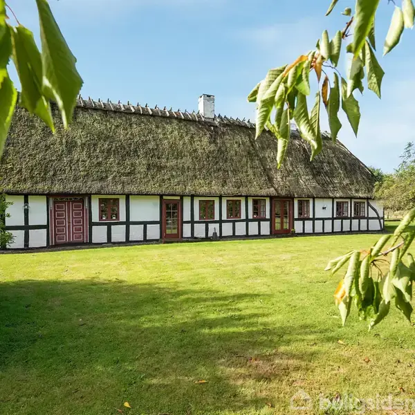 En traditionel bindingsværksbygning med stråtag står stille i en grøn have. Træer og blå himmel omkranser huset, hvilket skaber en fredfyldt atmosfære.