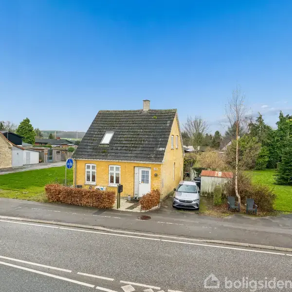 Et lille gult hus står stille ved en vejkant, med en parkeret bil og grønne træer i baggrunden under en klar blå himmel.