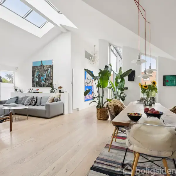 Stue med moderne indretning; en grå sofa og træstole i venstre side, spisebord med farverige stole til højre. Store vinduer og planter skaber en lys og åben atmosfære.