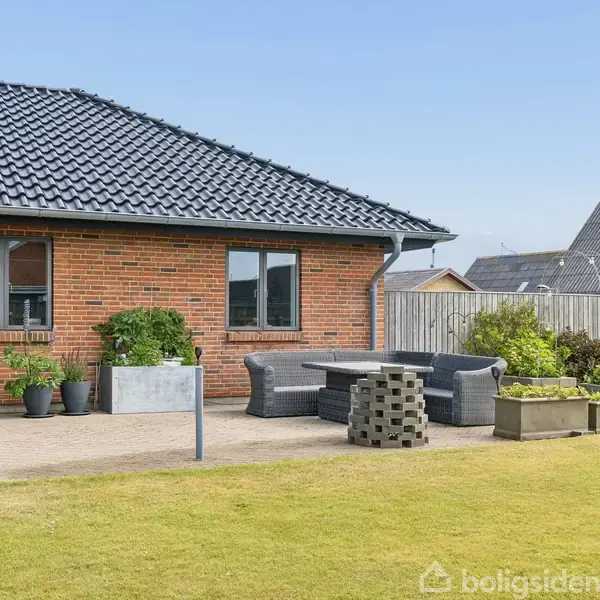 Rødt murstenshus med sorte tagsten, står i en have. På terrassen står et sæt grå udendørsmøbler og flere planter i potter. Baggrunden viser andre huse og en blå himmel.