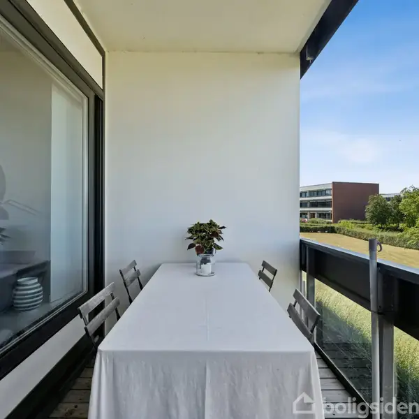 En balkon med et bord dækket med hvid dug står omgivet af stole. En plante pynter bordet. I baggrunden ses græsarealer og moderne bygninger under en klar blå himmel.