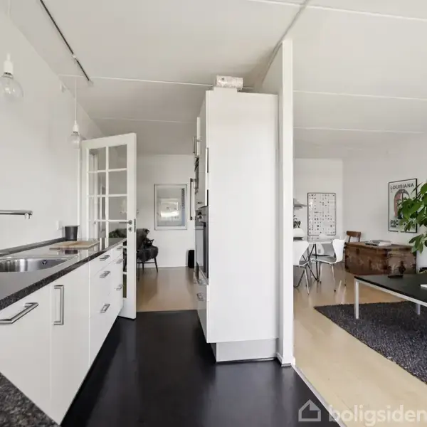 Et moderne køkken med hvide skabe og en sort bordplade strækker sig langs en væg. Åben forbindelse til en stue med et bord, en stor plante og lyse trægulve.