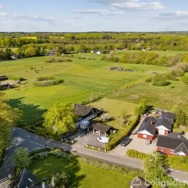 Et landligt område med flere bygninger omgivet af marker og skov. En vej løber langs ejendommene, som består af både røde og mørke bygninger. Skyfri himmel og frodig natur i baggrunden.
