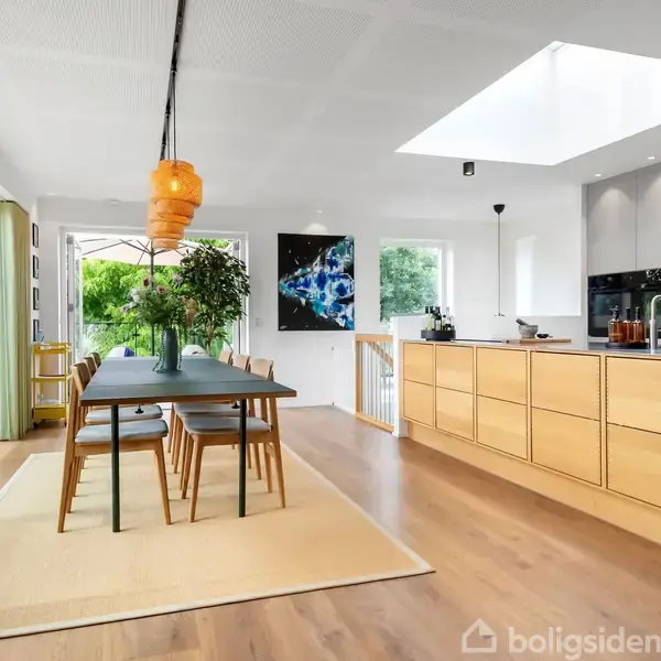 Et langbord med seks stole står i et lyst, moderne køkken-alrum. Et stort ovenlys og glasdør giver naturligt lys. Plante og kunst dekorerer rummet, omgivet af lyse træoverflader.