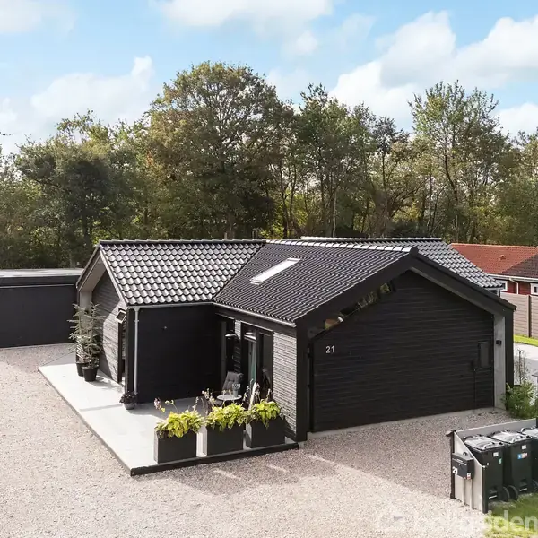 Et sortmalet hus står stille på en grusindkørsel omkranset af grønne buske, med garage og terrasse; beliggende i et skovområde med andre boliger. Husnummer "21" er synligt.