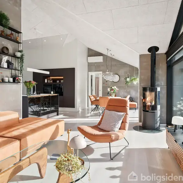 En stue med moderne møbler, herunder en orange lænestol og sofa, står i et lyst rum med store vinduer og en pejs. Et spisebord og køkken ses i baggrunden.
