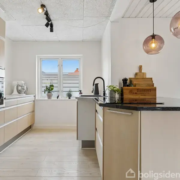 Et moderne køkken med lyse træskabe og sorte bordplader. To runde lamper hænger over en køkkenø. En vindueskarm har planter. Et skærebræt og en mixer ses på bordene.
