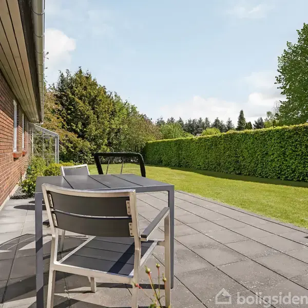 Terrassemøbler står på en flisebelagt terrasse, omkranset af en velholdt grøn hæk og græsplæne. En rød murstensmur til venstre indrammer området under en klar blå himmel.