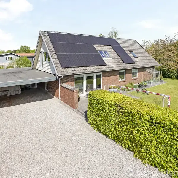 Et hus med solpaneler på taget står ved siden af en grusindkørsel. En velplejet have med græs og buske omgiver huset. Carporten er placeret til venstre.
