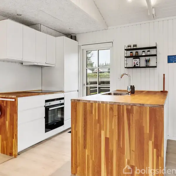 Køkken med træbordplader og hvide skabe. En dør fører ud til haven, og der er hylder med krus og en klock. Skilt: "Every day is a fresh start."