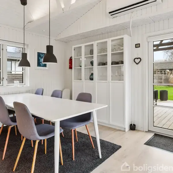 Et spisebord med seks stole står på et tæppe, mens to lamper hænger over bordet. Rummet har hvide vægge, et vitrineskab og en glasdør, som fører ud til en terrasse.