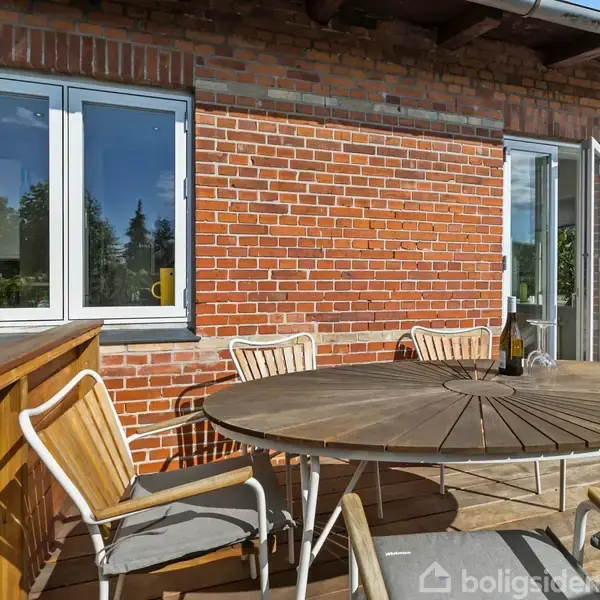 Rundt havebord med træstole står på en terrasse foran en murstensvæg. I baggrunden ses et vindue og en glasdør, som fører til indendørsarealet.