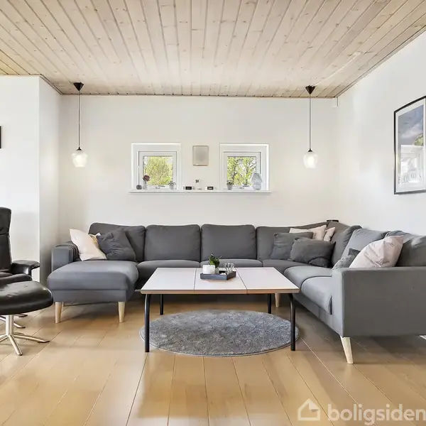 Gråt hjørnesofa står i en stue med lyse trægulve og loft. To små vinduer bagved, hængende lamper og et sort læderlænestol med fodskammel ved siden af.