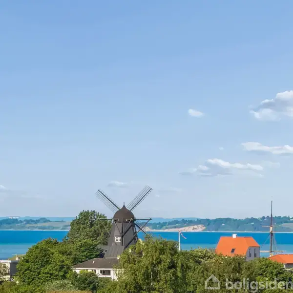En vindmølle står stationær i et naturskønt område med udsigt over vandet og fjernliggende land i baggrunden. Vegetation og bygninger omkranser møllen under en klar blå himmel.