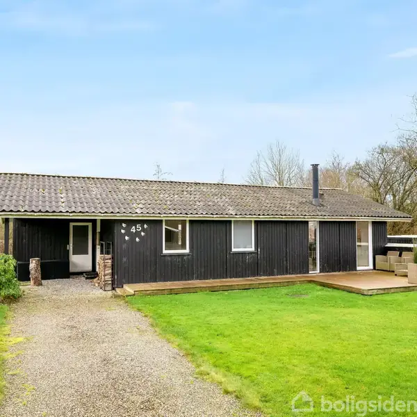 Et sort, enkelt etplanshus med nummer 45 ved hoveddøren står i en grøn have. En grussti leder op til huset, og der er en terrasse med havemøbler. Skov i baggrunden.