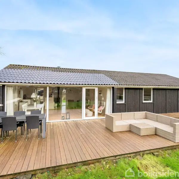 En moderne, sortmalet villa med store glaspartier åbner ud mod en rummelig træterrasse. Terrassen er indrettet med spisebord og loungeområde, omgivet af en grøn have.