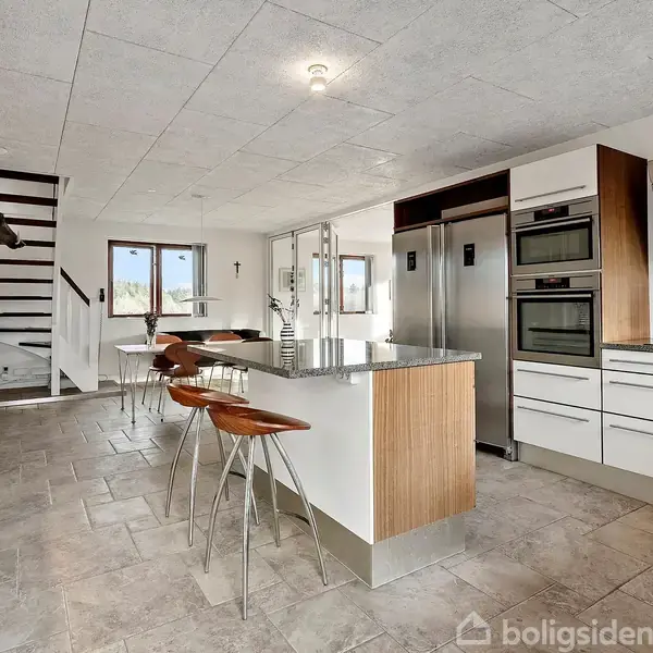 Køkken med moderne hvide skabe og grå bordplader, hvor en køkkenø har tre brune barstole. Trappe fører til øvre etage. Stort vindue til venstre giver naturligt lys.