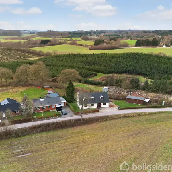 To huse ligger ved en vej. Omkring dem er store marker, tæt skov og bakker. Mange træer ses i baggrunden under en blå himmel med få skyer.