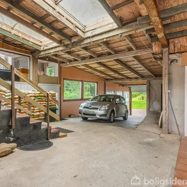 Bilen står parkeret i en rummelig garage med rå trækonstruktioner og åbent loft. Trætrapper fører til en anden etage, og store vinduer viser en grøn udendørs omgivelser.