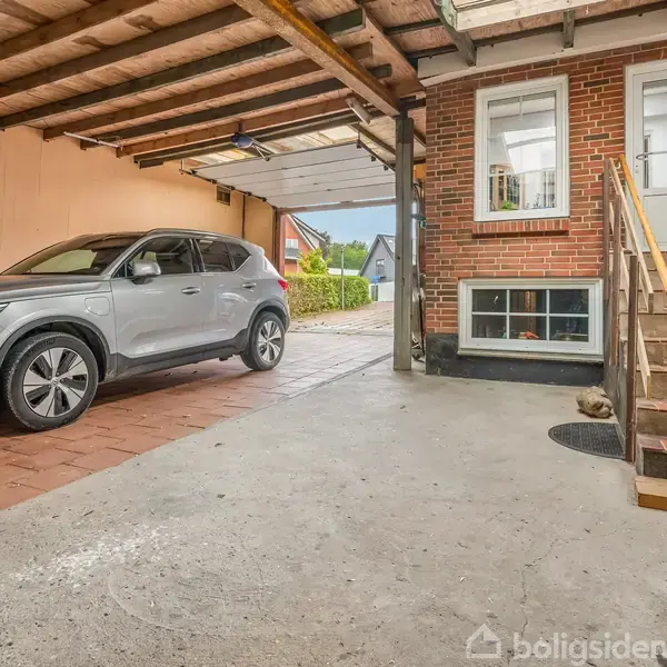 En sølvfarvet SUV er parkeret i en carport ved siden af et murstenshus med hvidt dør- og vinduesrammer. Trappe fører op til husets indgang.