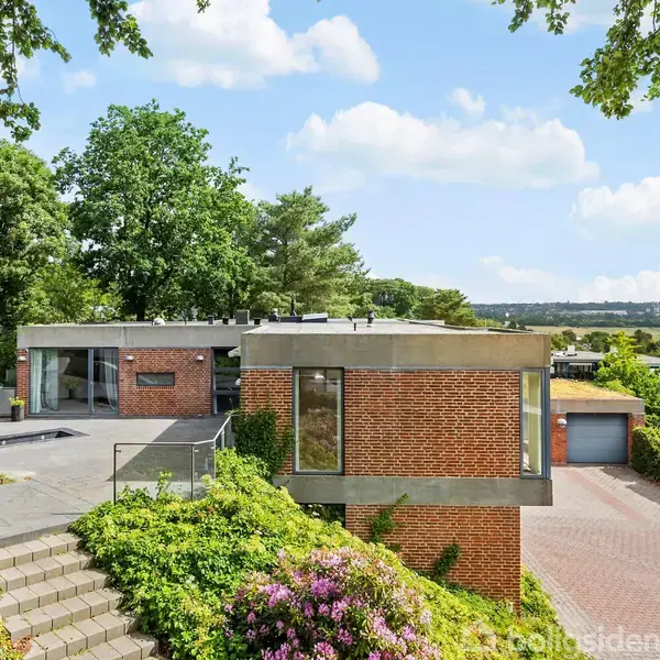 Et moderne hus med murstensvægge omgivet af grønne træer og buske. Trapper fører til indgangen. I baggrunden ses en udsigt over et grønt landskab og en garage.