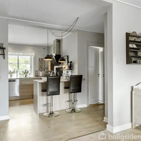 Et køkkenområde med en ø, hvor to høje stole står ved en bardisk. Omgivelserne er lyst indrettet med lyse træmøbler og hvide vægge.