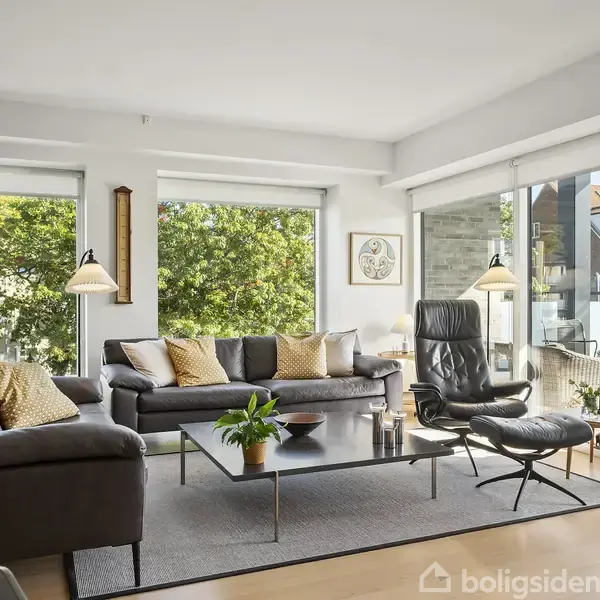 Stue med grå sofaer og sort lænestol omkring et sofabord med planter. Store vinduer giver udsigt til en terrasse og træer udenfor, der skaber en lys, luftig atmosfære.