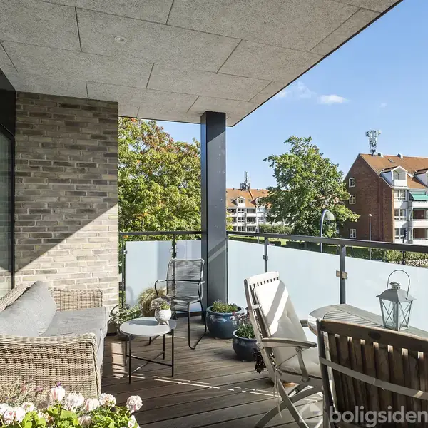 En udendørs altan med kurvemøbler og blomster i krukker. Den er delvist overdækket, og omgivet af glasrækværk. I baggrunden ses et boligområde med flerfamiliehuse og træer.